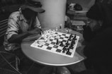 Two people are seated at a small round table, intensely focused on a game of chess. They are surrounded by chairs and other items. One person wears a hat and light clothing, while the other is dressed in darker attire and glasses. The image is in black and white.
