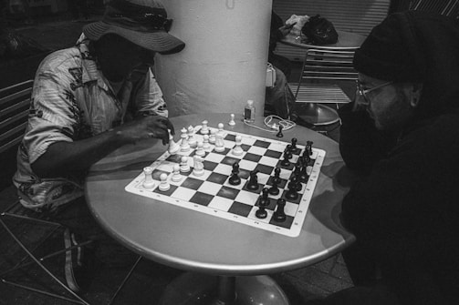 Two people are seated at a small round table, intensely focused on a game of chess. They are surrounded by chairs and other items. One person wears a hat and light clothing, while the other is dressed in darker attire and glasses. The image is in black and white.
