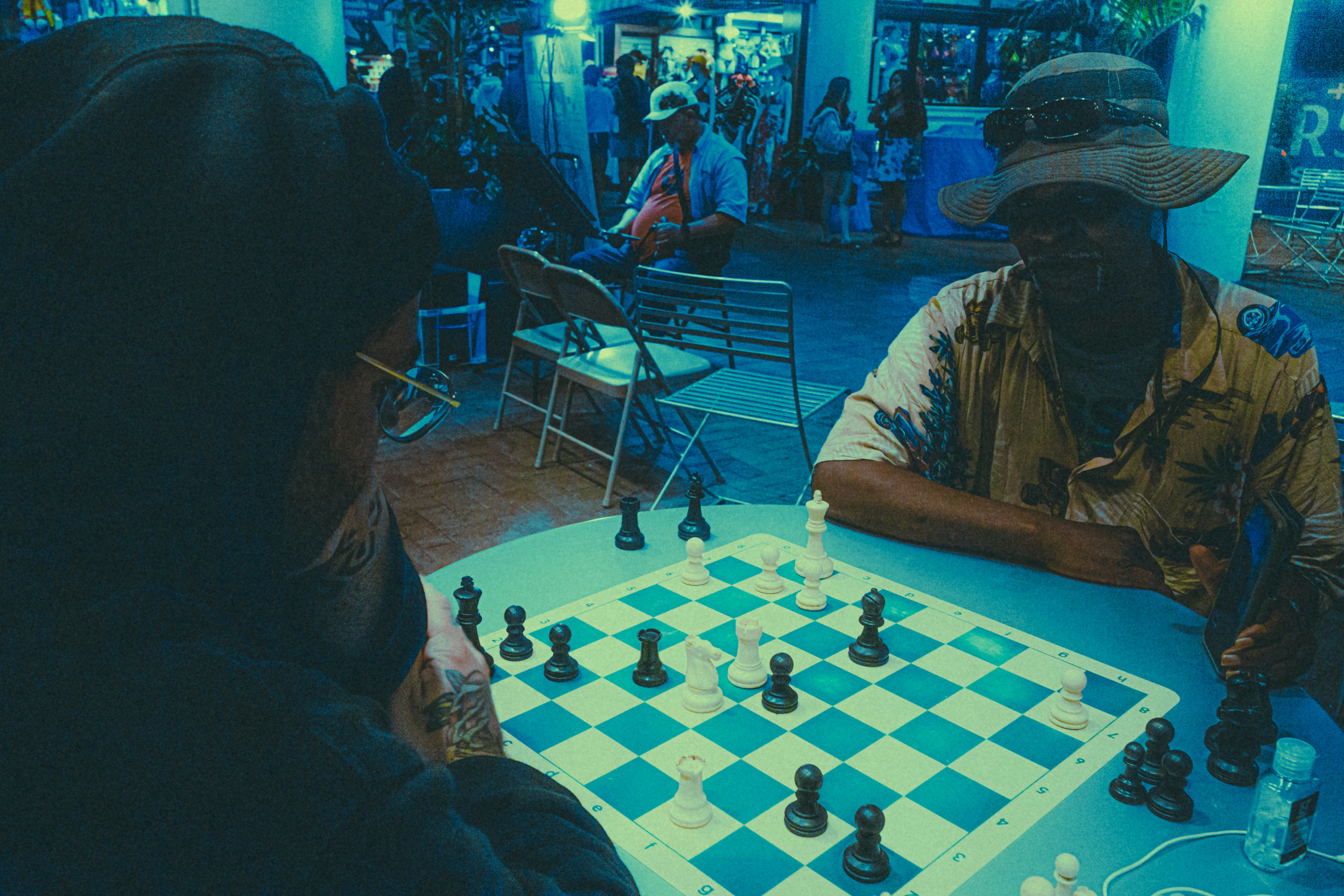 Two individuals are engaged in an intense game of chess on a small table. The scene is set in a dimly lit area with a bluish tint, possibly in an indoor or semi-outdoor location with chairs and other people in the background. One player is wearing a hoodie and glasses, while the other sports a wide-brimmed hat and a patterned shirt. The chessboard is prominently featured with pieces in mid-game, indicating active participation.