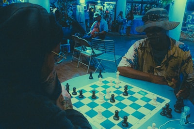 Two individuals are engaged in an intense game of chess on a small table. The scene is set in a dimly lit area with a bluish tint, possibly in an indoor or semi-outdoor location with chairs and other people in the background. One player is wearing a hoodie and glasses, while the other sports a wide-brimmed hat and a patterned shirt. The chessboard is prominently featured with pieces in mid-game, indicating active participation.