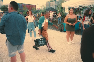 People gather in an outdoor market setting, featuring individuals casually dressed and engaging with one another. A man wearing striped pants and overalls carries a crate, while other people stand and converse near stalls and tables adorned with decorations. The background includes colorful murals and urban buildings, creating a lively atmosphere.