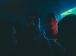 A dimly lit party scene with several people in close proximity, illuminated by blue and red neon lights. One person is holding a red plastic cup, giving the impression of a festive or club atmosphere.