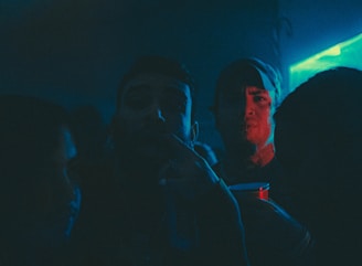 A dimly lit party scene with several people in close proximity, illuminated by blue and red neon lights. One person is holding a red plastic cup, giving the impression of a festive or club atmosphere.