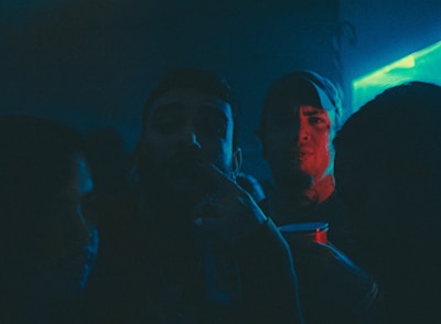 A dimly lit party scene with several people in close proximity, illuminated by blue and red neon lights. One person is holding a red plastic cup, giving the impression of a festive or club atmosphere.