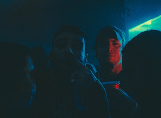 A dimly lit party scene with several people in close proximity, illuminated by blue and red neon lights. One person is holding a red plastic cup, giving the impression of a festive or club atmosphere.