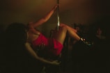 A person wearing a red dress is performing on a dance pole in a dimly lit environment surrounded by people. The scene has a moody, intimate atmosphere with low visibility due to darkness.
