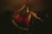 A person wearing a red dress is performing on a dance pole in a dimly lit environment surrounded by people. The scene has a moody, intimate atmosphere with low visibility due to darkness.