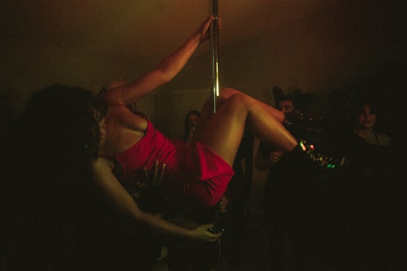 A person wearing a red dress is performing on a dance pole in a dimly lit environment surrounded by people. The scene has a moody, intimate atmosphere with low visibility due to darkness.