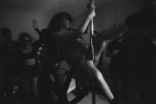 A dimly lit room filled with people and a central figure elegantly performing pole dancing. The crowd appears engaged and the setting indicates a lively, energetic atmosphere. The image has a grainy, monochrome aesthetic contributing to its moody ambiance.