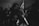 A dimly lit room filled with people and a central figure elegantly performing pole dancing. The crowd appears engaged and the setting indicates a lively, energetic atmosphere. The image has a grainy, monochrome aesthetic contributing to its moody ambiance.
