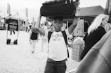 A black and white photograph of a young boy carrying a suitcase on his head while walking through a bustling outdoor market. He is wearing a sleeveless shirt with the text 'Levi Strauss and Co.' The background contains market stalls, people browsing, and a sign that reads 'Everyone from everywhere is welcome here'. The atmosphere seems lively with various stalls and people.