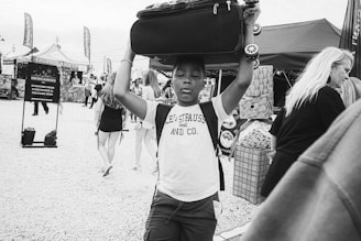A black and white photograph of a young boy carrying a suitcase on his head while walking through a bustling outdoor market. He is wearing a sleeveless shirt with the text 'Levi Strauss and Co.' The background contains market stalls, people browsing, and a sign that reads 'Everyone from everywhere is welcome here'. The atmosphere seems lively with various stalls and people.