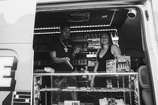 Two people are inside a van converted into a mobile store, displaying various products such as snacks and smoking-related items. The front counter is glass, showcasing merchandise for sale. The interior is well-lit, highlighting shelves filled with products. One person appears to be interacting or transacting with the other, suggesting a business or customer service interaction.