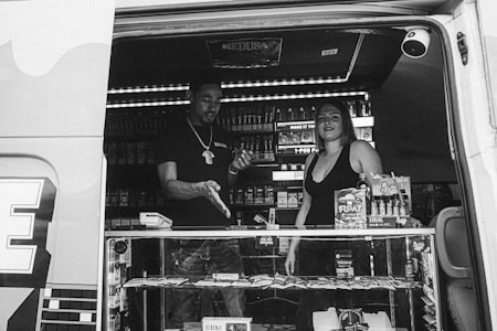 Two people are inside a van converted into a mobile store, displaying various products such as snacks and smoking-related items. The front counter is glass, showcasing merchandise for sale. The interior is well-lit, highlighting shelves filled with products. One person appears to be interacting or transacting with the other, suggesting a business or customer service interaction.