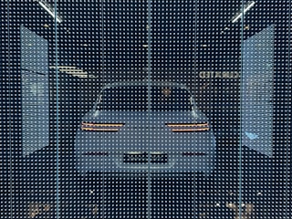 A white car displayed behind a glass panel covered in a matrix of small, bright LED lights creating a futuristic ambiance. The car's rear lights are distinctly visible, surrounded by a dark background that emphasizes the glowing pattern.