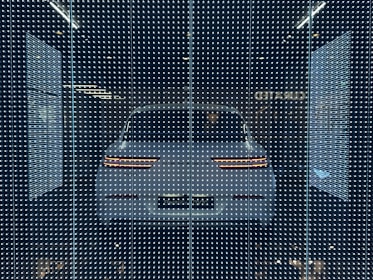 A white car displayed behind a glass panel covered in a matrix of small, bright LED lights creating a futuristic ambiance. The car's rear lights are distinctly visible, surrounded by a dark background that emphasizes the glowing pattern.