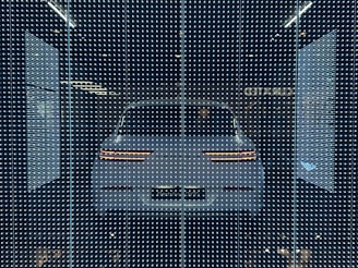 A white car displayed behind a glass panel covered in a matrix of small, bright LED lights creating a futuristic ambiance. The car's rear lights are distinctly visible, surrounded by a dark background that emphasizes the glowing pattern.