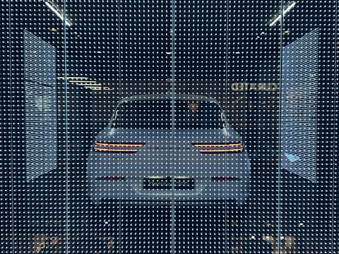 A white car displayed behind a glass panel covered in a matrix of small, bright LED lights creating a futuristic ambiance. The car's rear lights are distinctly visible, surrounded by a dark background that emphasizes the glowing pattern.
