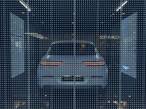 A white car displayed behind a glass panel covered in a matrix of small, bright LED lights creating a futuristic ambiance. The car's rear lights are distinctly visible, surrounded by a dark background that emphasizes the glowing pattern.