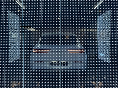 A white car displayed behind a glass panel covered in a matrix of small, bright LED lights creating a futuristic ambiance. The car's rear lights are distinctly visible, surrounded by a dark background that emphasizes the glowing pattern.
