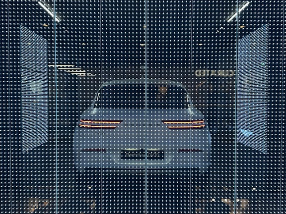 A white car displayed behind a glass panel covered in a matrix of small, bright LED lights creating a futuristic ambiance. The car's rear lights are distinctly visible, surrounded by a dark background that emphasizes the glowing pattern.