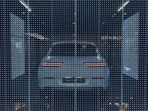 A white car displayed behind a glass panel covered in a matrix of small, bright LED lights creating a futuristic ambiance. The car's rear lights are distinctly visible, surrounded by a dark background that emphasizes the glowing pattern.