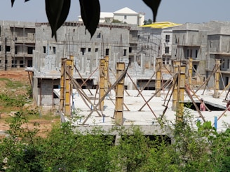 A construction site with partially built concrete structures and scaffolding in place. Several unfinished buildings are visible, as well as vegetation in the foreground. A worker wearing a hard hat is seen at the site.