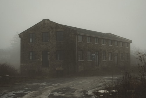 A large, old stone building stands in a misty setting. The structure has numerous rectangular windows and appears to be abandoned. Overgrowth surrounds the building and the ground is uneven, suggesting neglect. The overall atmosphere is eerie and atmospheric.