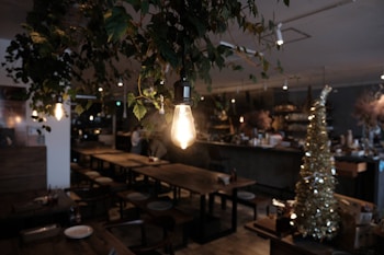 A cozy cafe interior with dim lighting, featuring an exposed light bulb hanging from the ceiling surrounded by leafy plants. Wooden tables and chairs are arranged in an orderly fashion, with a decorated Christmas tree on the right side. The ambiance is warm and inviting, suitable for both social gatherings and quiet moments.