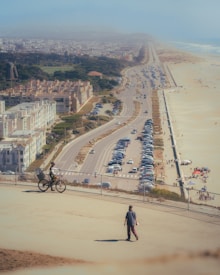 A scenic coastal view with a long road running parallel to the beach. The area is populated with buildings on one side and the ocean on the other. A cyclist is riding along a path with a person walking nearby. The beach has small groups of people under umbrellas, and many cars are parked along the road.