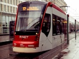 A modern red and white tram is stationed on a tramway in an urban environment. Reflections of rain can be seen on the wet pavement, and the illuminated destination sign on the tram reads 'Den Haag Kunstmuseum'. The surrounding area features contemporary buildings with glass and metal structures.