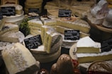 Various types of cheese are displayed on a counter, each labeled with tags that include their names and prices. The cheeses are of different shapes, some round and some cut into wedges, showcasing textures like creamy interiors and moldy rinds.