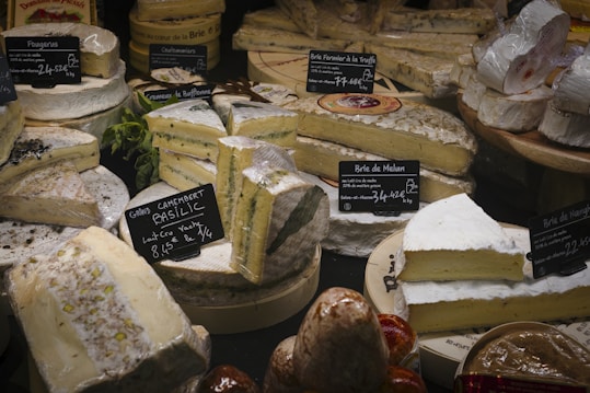 Various types of cheese are displayed on a counter, each labeled with tags that include their names and prices. The cheeses are of different shapes, some round and some cut into wedges, showcasing textures like creamy interiors and moldy rinds.