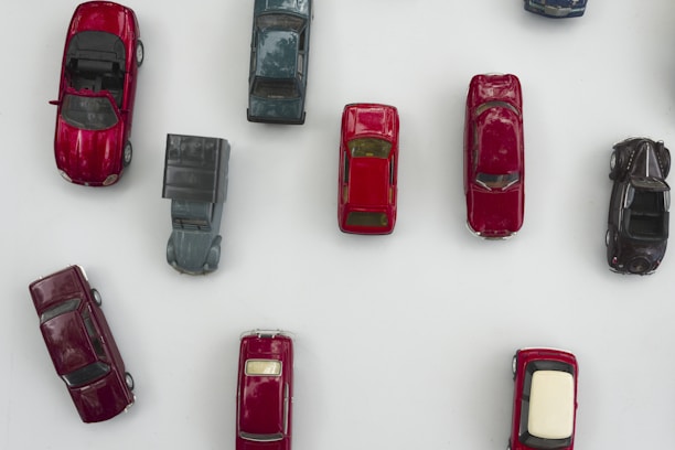 A collection of miniature toy cars arranged on a white surface. The toys are of various colors and types, including vintage and modern designs. The vehicles are mostly in shades of red and black, with one older, boxier car standing out in a muted gray.
