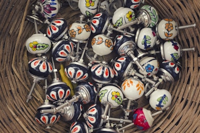 A collection of colorful ceramic drawer knobs with floral and geometric patterns, placed inside a woven basket. The knobs have metal screws attached, and they feature designs in a variety of colors including red, blue, yellow, and green.