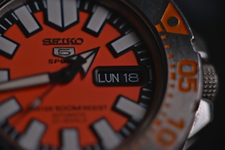 A close-up of a sports wristwatch with an orange and black face, featuring bold white hour markers and a date window displaying 'LUN 18'. The watch hand is prominently visible, and there is text indicating 'SEIKO 5 Sports' along with additional text in smaller font.