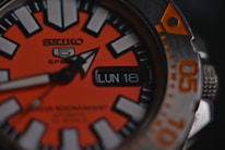 A close-up of a sports wristwatch with an orange and black face, featuring bold white hour markers and a date window displaying 'LUN 18'. The watch hand is prominently visible, and there is text indicating 'SEIKO 5 Sports' along with additional text in smaller font.