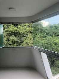 A balcony with a clear glass railing provides an unobstructed view of dense, lush green foliage. The balcony's floor and walls are light gray, and the overcast sky adds a soft light to the scene.