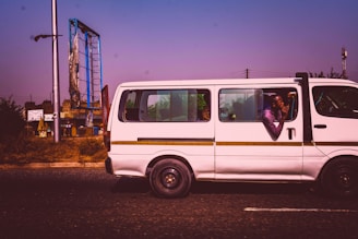 A white minibus is traveling on a road with a clear purple sky above. A man inside the bus leans out the window, appearing relaxed, with his elbows resting on the window edge. In the background, there is a damaged billboard supported by a tall metal frame, and some green foliage is visible.