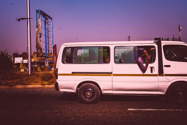 A white minibus is traveling on a road with a clear purple sky above. A man inside the bus leans out the window, appearing relaxed, with his elbows resting on the window edge. In the background, there is a damaged billboard supported by a tall metal frame, and some green foliage is visible.