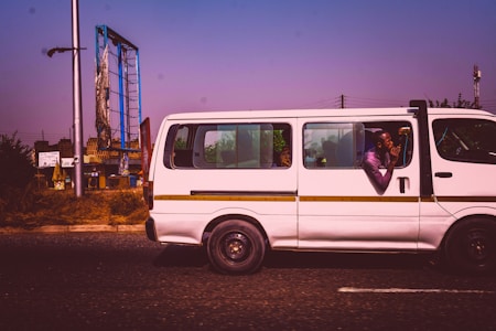 A white minibus is traveling on a road with a clear purple sky above. A man inside the bus leans out the window, appearing relaxed, with his elbows resting on the window edge. In the background, there is a damaged billboard supported by a tall metal frame, and some green foliage is visible.
