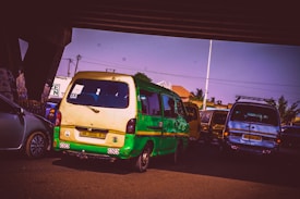 A scene under a bridge features several vehicles, including a prominently placed green and yellow minibus with visible scratches on its body. The surroundings include urban elements like buildings and trees, visible in the background against a clear sky.