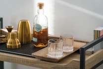 A wooden cart holds a bottle of beverage labeled 'Roe & Co Dublin', alongside two empty glass tumblers placed on a rectangular wooden tray. The cart also contains a set of golden cocktail shakers and jiggers. In the background, there is a stack of colorful small books or boxes.