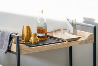 A well-organized bar cart displays an assortment of cocktail-making tools and a bottle of whiskey. Two gold-colored cocktail shakers, a gold jigger, and a strainer are neatly arranged on a black mat. A towel is draped over the edge of the cart. Two empty glass tumblers and a set of colorful coasters are placed next to the whiskey bottle, which has a clear label.
