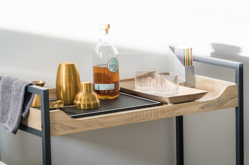 A well-organized bar cart displays an assortment of cocktail-making tools and a bottle of whiskey. Two gold-colored cocktail shakers, a gold jigger, and a strainer are neatly arranged on a black mat. A towel is draped over the edge of the cart. Two empty glass tumblers and a set of colorful coasters are placed next to the whiskey bottle, which has a clear label.