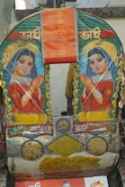 The image features a vibrant and ornately decorated rickshaw backplate. It includes artistic depictions of two women, both dressed in colorful traditional attire, with an expression of welcome. Surrounding the portraits are decorative elements such as patterns, flowers, and inscriptions in a script. The overall colors are vivid, including bright reds, greens, and yellows.