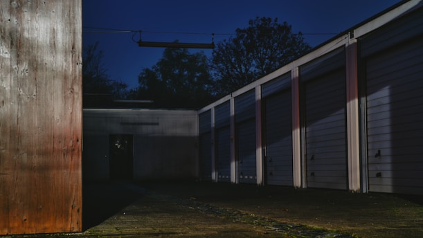A row of closed garage doors is aligned next to a wooden structure, casting long shadows on the ground. The scene is dark, with an ominous sky overhead and trees silhouetted in the background. The light creates a moody atmosphere, highlighting the textures of the wood and metal surfaces.