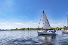 A sailboat with the name 'DIAMANT' glides across a calm body of water. The boat has a large white sail with a hint of blue on the edges and carries several people leisurely enjoying the day. In the background, lush green foliage lines the shore, with a few buildings visible in the distance under a clear blue sky.