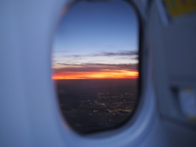 A vibrant sunset view captured from the window of an airplane, with a gradient of orange, red, and purple hues blending into the dark blue sky. The city lights below create a delicate tapestry, indicating urban areas beneath the flight path.