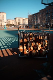 A cart filled with basketballs is positioned on an outdoor basketball court. The court is illuminated by sunlight, creating shadows and a vibrant atmosphere. In the background, several residential buildings surround the area, adding an urban feel to the setting.